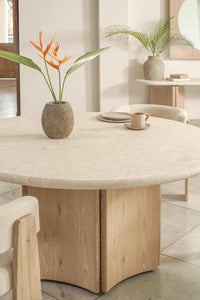 The Cesena | Dining Table with a light wood finish and sturdy wooden legs is styled with a stone vase holding a bird of paradise, palm leaf, cups, and plates. Neutral-toned chairs and minimalist decor complete the bright, modern room.