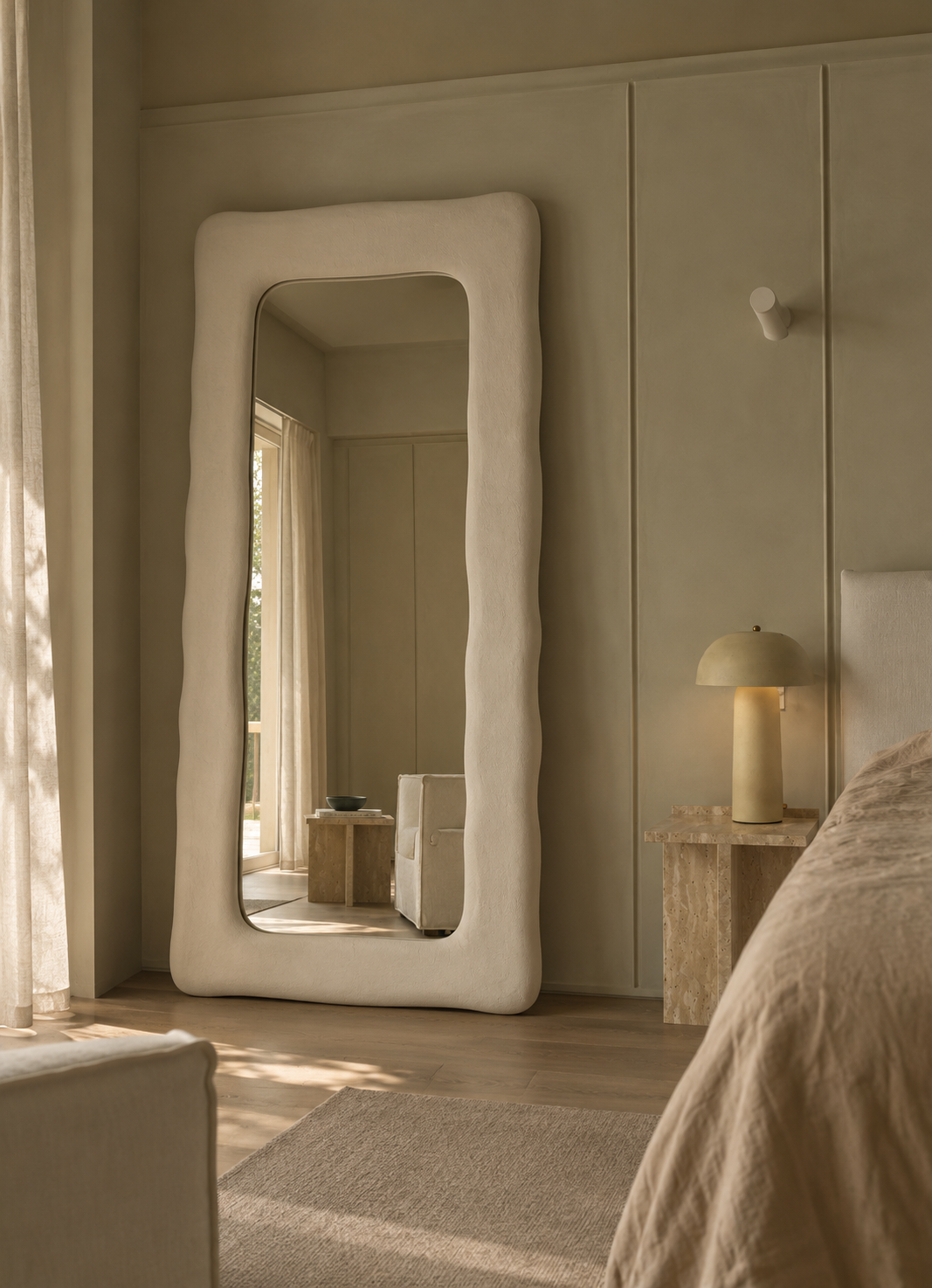 A tall floor mirror with a thick, wavy organic white frame stands in a softly lit bedroom. The mirror reflects part of the room, and the scene includes a bed, bedside lamp, curtains, and neutral-toned walls.