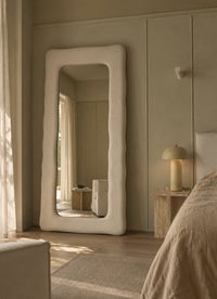 A tall floor mirror with a thick, wavy organic white frame stands in a softly lit bedroom. The mirror reflects part of the room, and the scene includes a bed, bedside lamp, curtains, and neutral-toned walls.