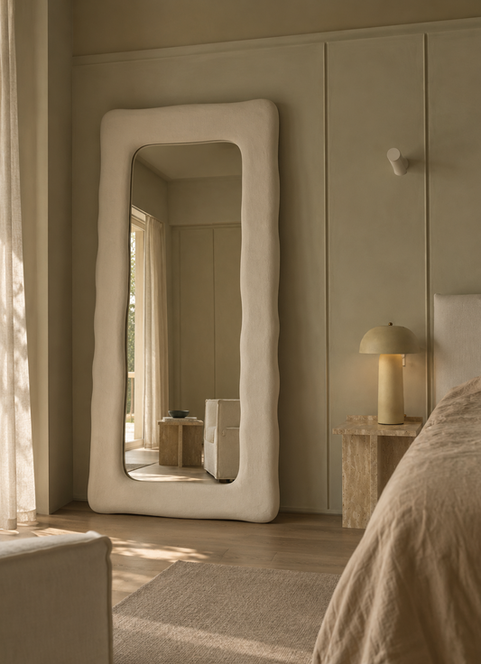 A tall floor mirror with a thick, wavy organic white frame stands in a softly lit bedroom. The mirror reflects part of the room, and the scene includes a bed, bedside lamp, curtains, and neutral-toned walls.