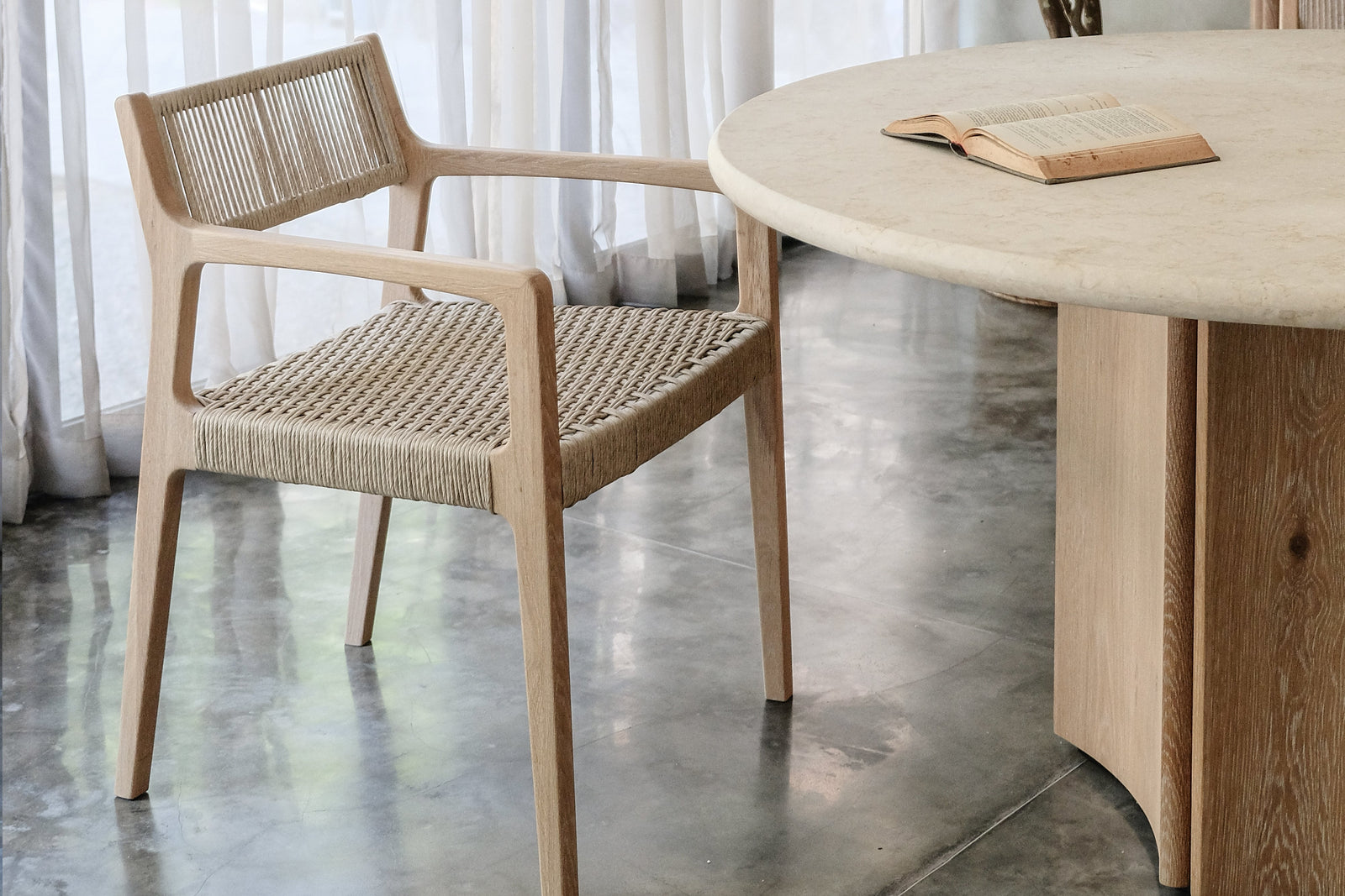 A wooden chair with a woven seat sits beside the Cesena | Dining Table, which has wooden legs and an open book on it. Sheer white curtains, a tall leafy plant, and polished concrete flooring complete the setting.