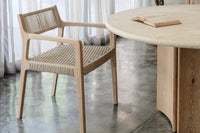 A wooden chair with a woven seat sits beside the Cesena | Dining Table, which has wooden legs and an open book on it. Sheer white curtains, a tall leafy plant, and polished concrete flooring complete the setting.