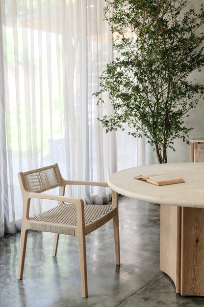 A wooden chair with a woven seat sits beside the Cesena | Dining Table, which has wooden legs and an open book on it. Sheer white curtains, a tall leafy plant, and polished concrete flooring complete the setting.