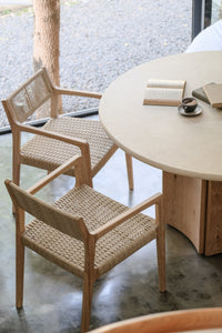 Two woven wooden chairs sit by the Cesena | Dining Table, which has wooden legs and a round marble top. On it are an open book, a closed book, and a cup of coffee. The table is set by a window overlooking trees and gravel outside.