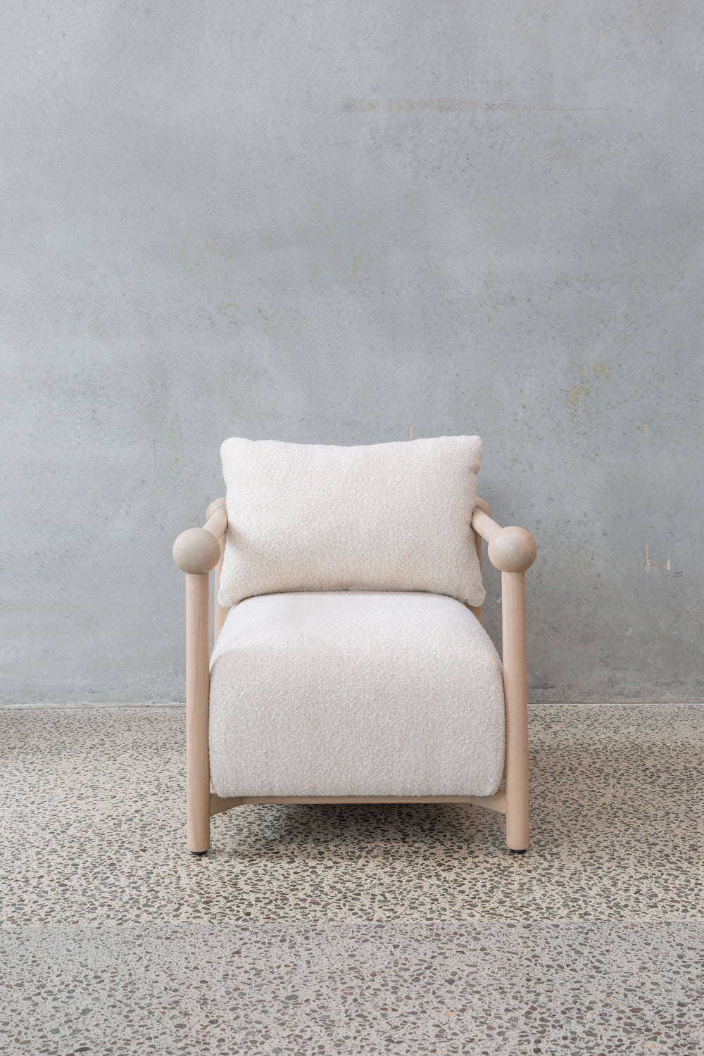 The Karia Armchair features a light wood frame, rounded armrests, and soft off-white upholstery, providing luxurious comfort on a speckled stone floor against a plain light gray wall.