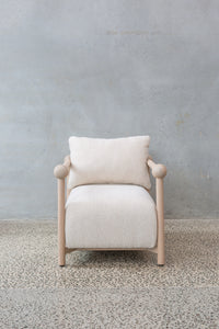 The Karia Armchair features a light wood frame, rounded armrests, and soft off-white upholstery, providing luxurious comfort on a speckled stone floor against a plain light gray wall.