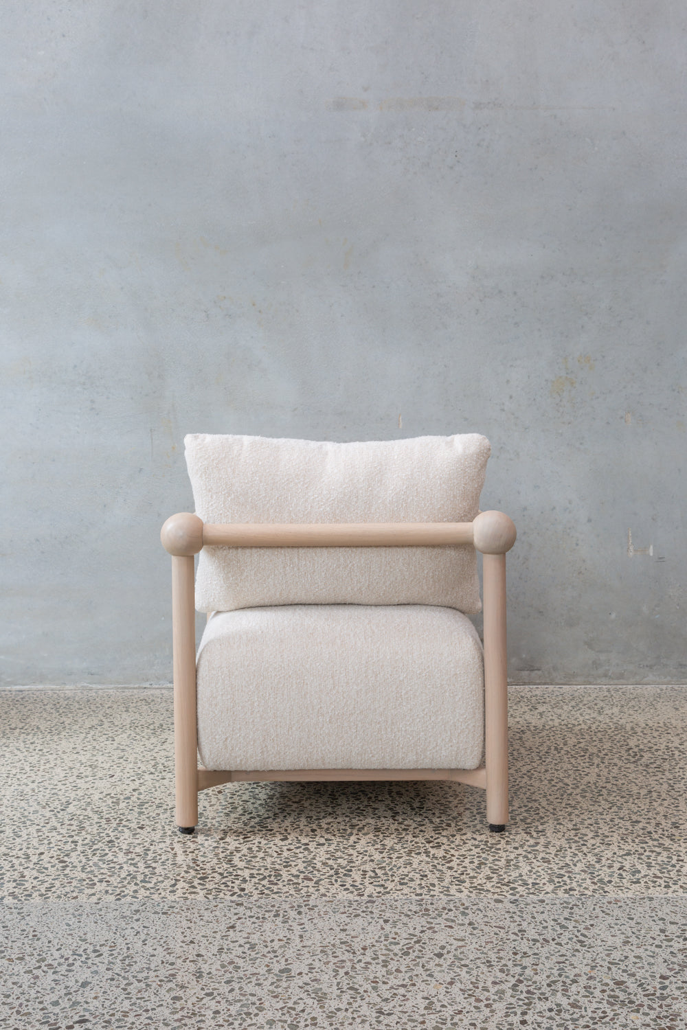 The Karia Armchair features a light wood frame and white cushion, offering comfortable seating on a speckled concrete floor with a plain, light gray textured wall background.