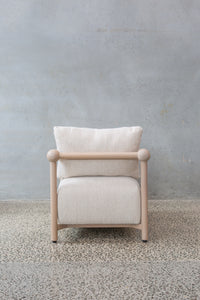 The Karia Armchair features a light wood frame and white cushion, offering comfortable seating on a speckled concrete floor with a plain, light gray textured wall background.