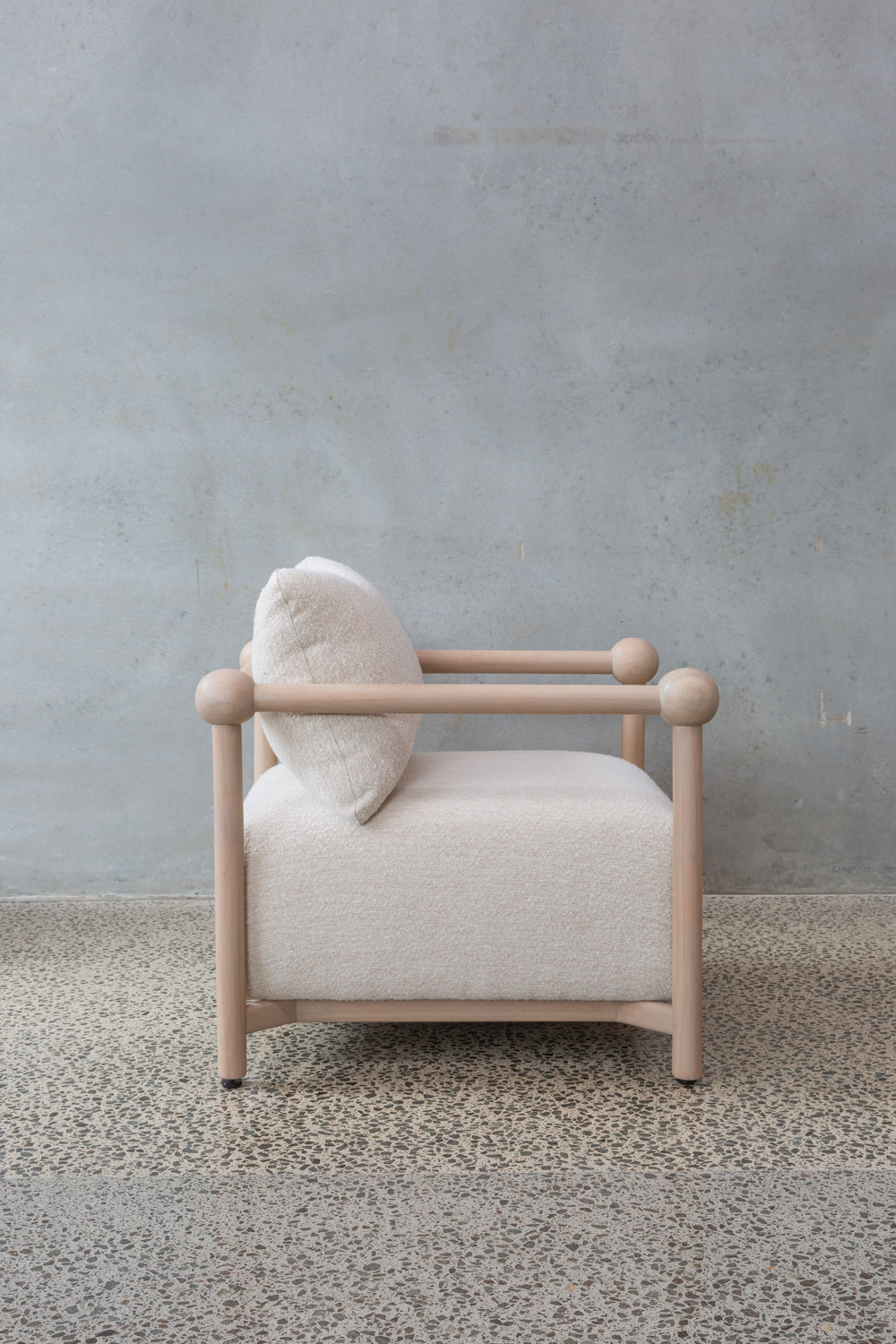 The Karia | Armchair features a light wooden frame, round armrests, and off-white cushioned seat and backrest, offering modern comfort on a speckled stone floor beside a textured gray wall.