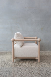 The Karia | Armchair features a light wooden frame, round armrests, and off-white cushioned seat and backrest, offering modern comfort on a speckled stone floor beside a textured gray wall.