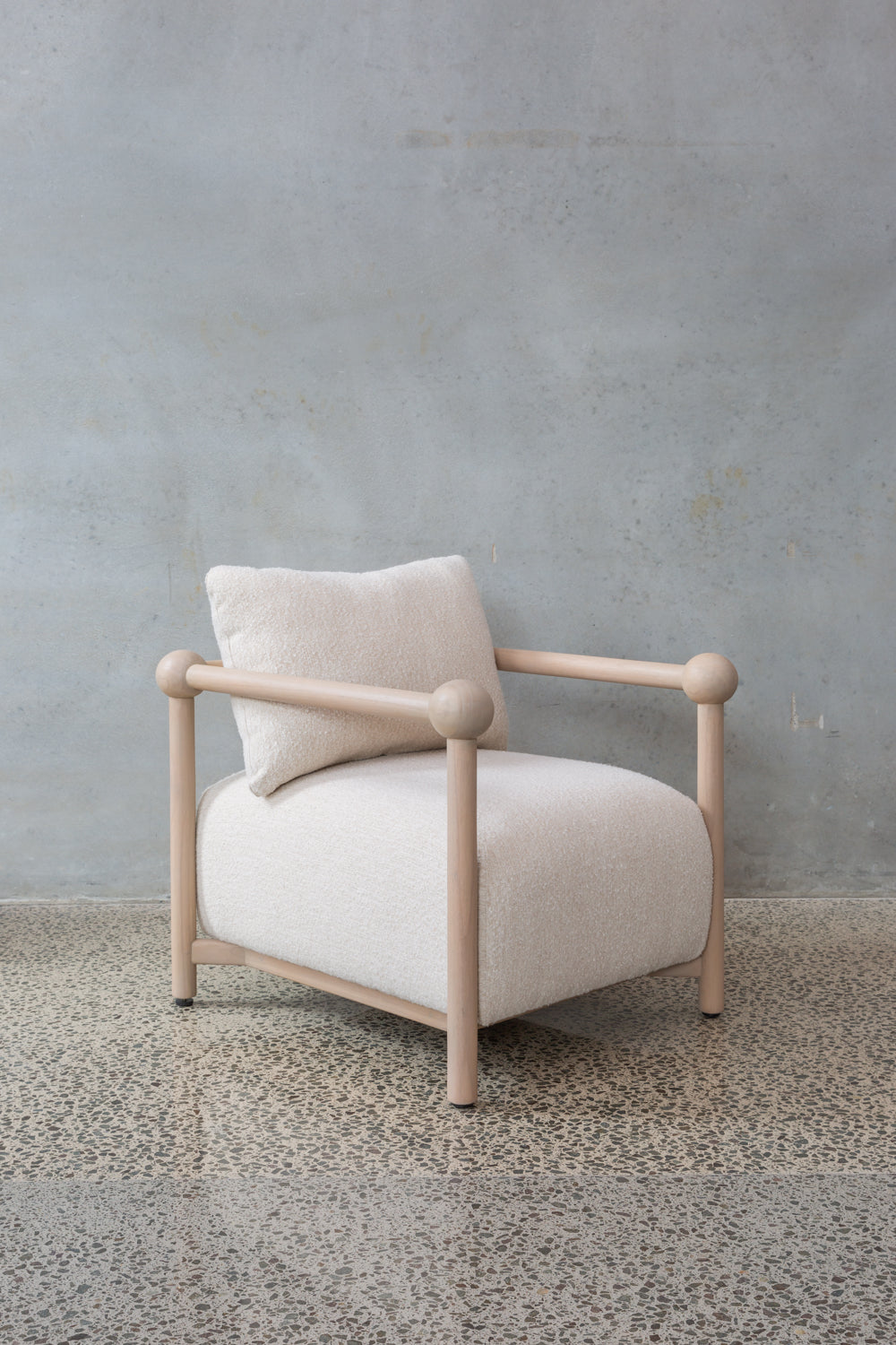 The Karia | Armchair features a light wood frame, rounded edges, and a textured off-white cushion and pillow, providing stylish seating atop a speckled stone floor with a gray wall backdrop.