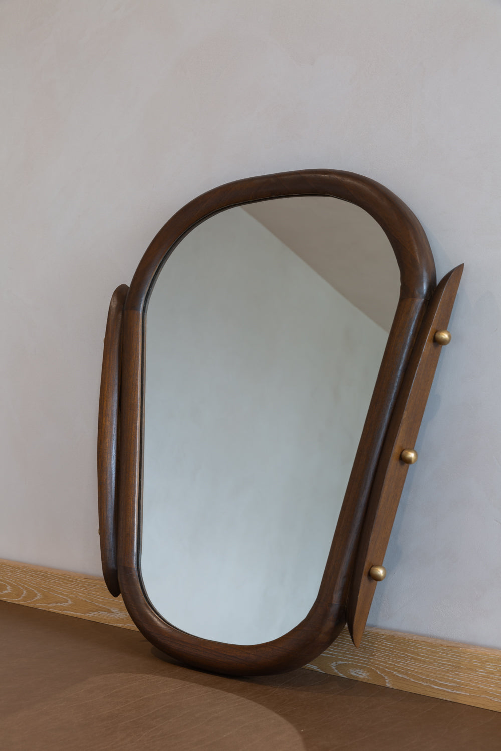 The Cedir | Mirror, featuring rounded edges and brass knobs, leans against a light wall atop wood. Its mid-century design reflects the room opposite, serving as a subtle yet stylish statement piece for any interior.