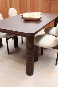 The Riad | Table features ribbed dark solid Mindi wood legs and is paired with four cream upholstered chairs. Two gold-toned decorative bowls sit atop the table, while light wood flooring and wood paneling enhance the modern style.
