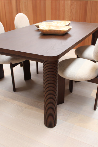 The Riad | Table features ribbed dark solid Mindi wood legs and is paired with four cream upholstered chairs. Two gold-toned decorative bowls sit atop the table, while light wood flooring and wood paneling enhance the modern style.