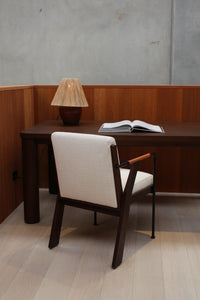 A minimalist workspace with a dark solid Mindi wood desk, open book, woven-shade lamp, and cushioned white chair on light floors reflects the refined elegance of the Riad | Table.