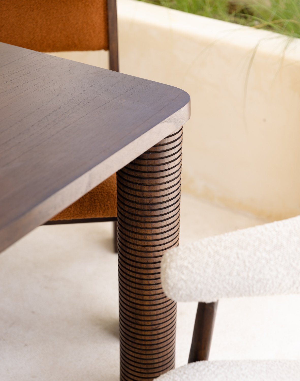 Close-up of the Riad | Table, a modern dining table made from solid Mindi wood with rounded edges and a ribbed cylindrical leg. A white textured chair and a brown-cushioned chair are placed nearby, set against a light wall and green plants.