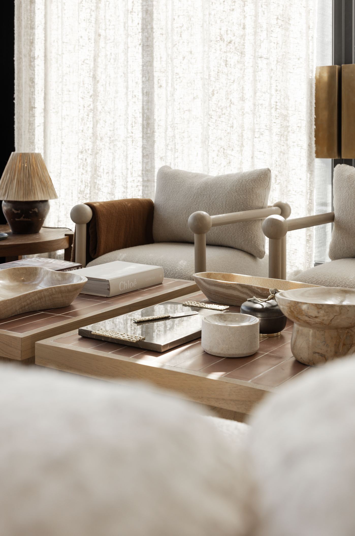 A cozy, modern living room featuring Karia | Armchair in neutral tones, textured cushions, and wooden coffee tables adorned with decorative bowls, books, and trays—all illuminated by soft natural light through sheer curtains.