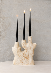 A three-arm candle holder made of natural beige stone with black taper candles, shown lit on a light surface against a textured gray wall.
